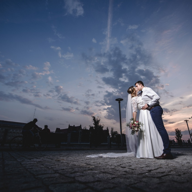 Picturesque wedding photoshoot with grey skies at dusk, bride and groom celebrating with a romantic kiss, groom has removed his coat