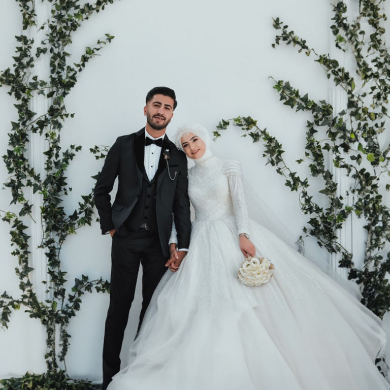 Wedding photoshoot with the bride and groom standing side by side, holding hands, white background with ivy on the walls