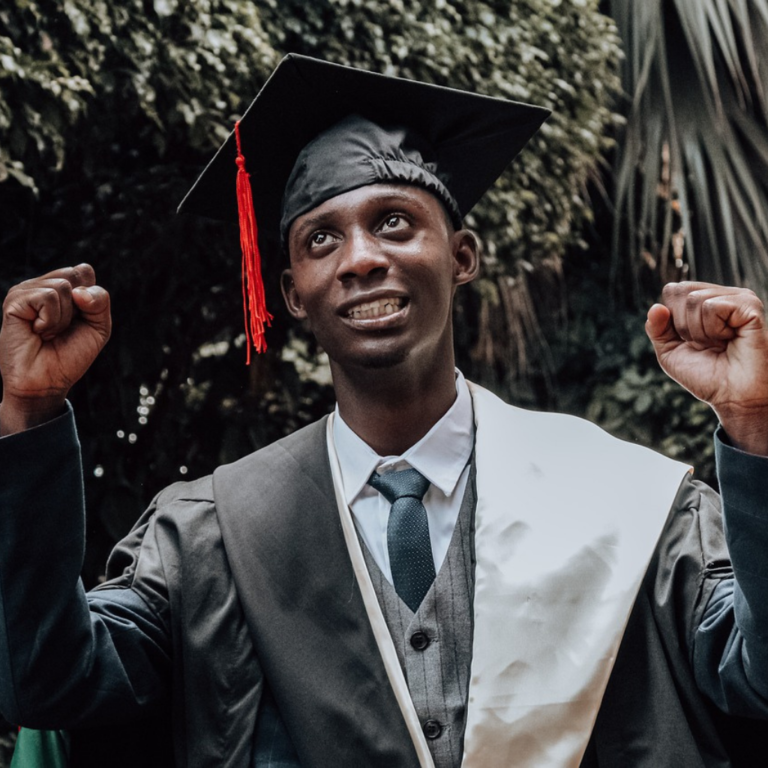 Graduation photoshoot, the graduand is wearing a black graduate cap, a suit and a tie, holding his hands up as a mark of success and achievement