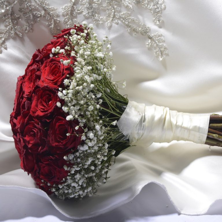 Wedding bouquet of red roses, and other flower arrangements, tied with white satin laid on a white wedding gown as background