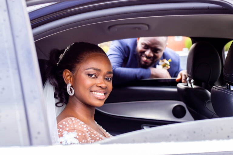 Wedding Photoshoot by Quintessence Photography UK. The bride smiling in the car as the groom watches from behind